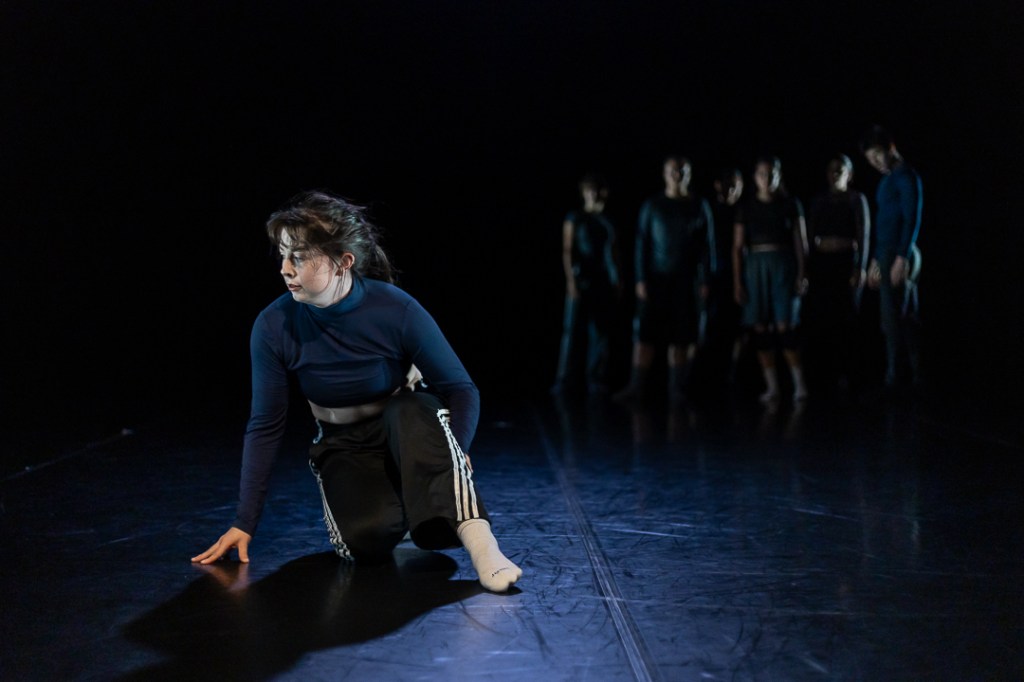 dancer crouched downstage in focus with group clustered upstage